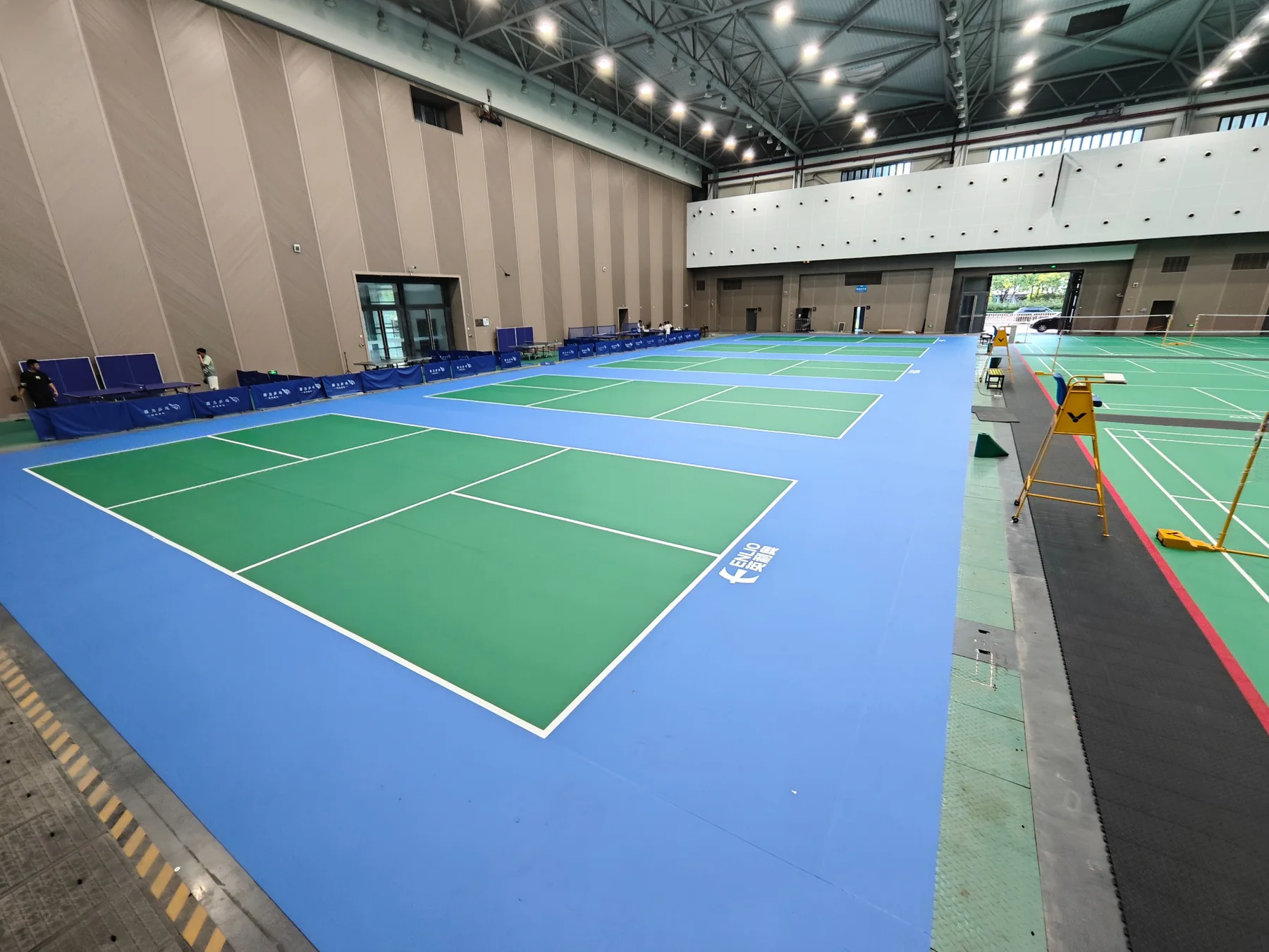 Large indoor hall with multiple green pickleball courts and blue surrounds
